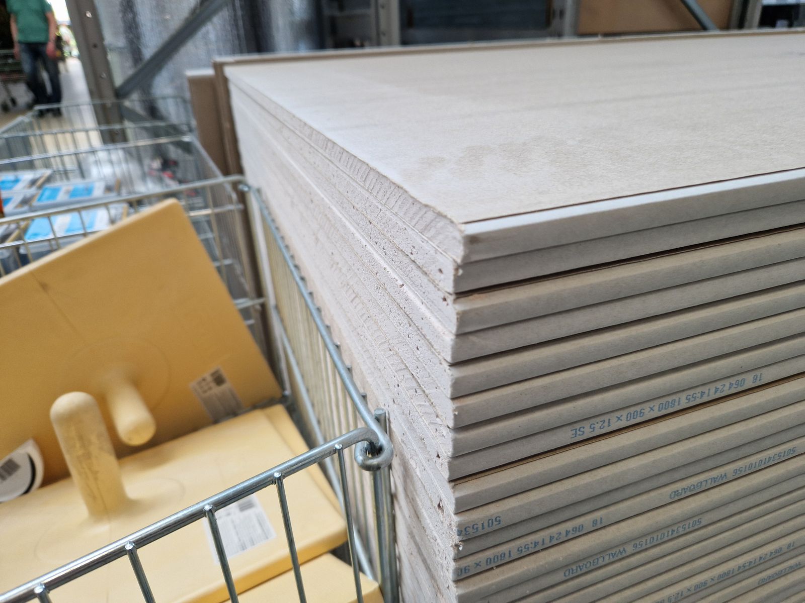 A stack of grey plasterboard / wallboard sheets on a wire stillage in a store, the visible edges printed with generic size and spec text, with a wire trolley of other stock alongside.